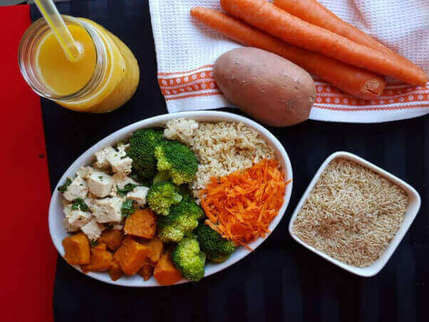 a vegan macro bowl made with tofu, broccoli, brown rice, sweet potatoes, and carrots on a table. There is a glass of orange juice next to the bowl