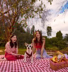 Sisters Violet and Madeleine McGraw sit on a red and white picnic blanket outside on the grass with their dog Rudy