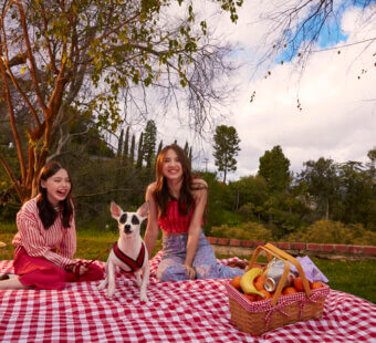 Sisters Violet and Madeleine McGraw sit on a red and white picnic blanket outside on the grass with their dog Rudy