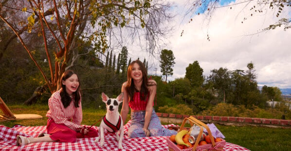 Sisters Violet and Madeleine McGraw sit on a red and white picnic blanket outside on the grass with their dog Rudy