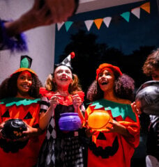 four kids dressed up for Halloween at a neighbor's door asking for candy. It is dark outside and two of the kids are dressed up as pumpkins, one kid is dressed up as a clown, and the other kid is dressed up as a skeleton.