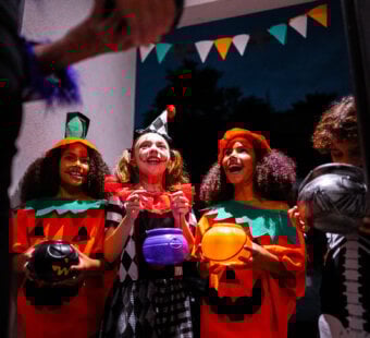 four kids dressed up for Halloween at a neighbor's door asking for candy. It is dark outside and two of the kids are dressed up as pumpkins, one kid is dressed up as a clown, and the other kid is dressed up as a skeleton.