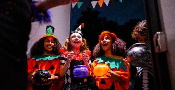four kids dressed up for Halloween at a neighbor's door asking for candy. It is dark outside and two of the kids are dressed up as pumpkins, one kid is dressed up as a clown, and the other kid is dressed up as a skeleton.