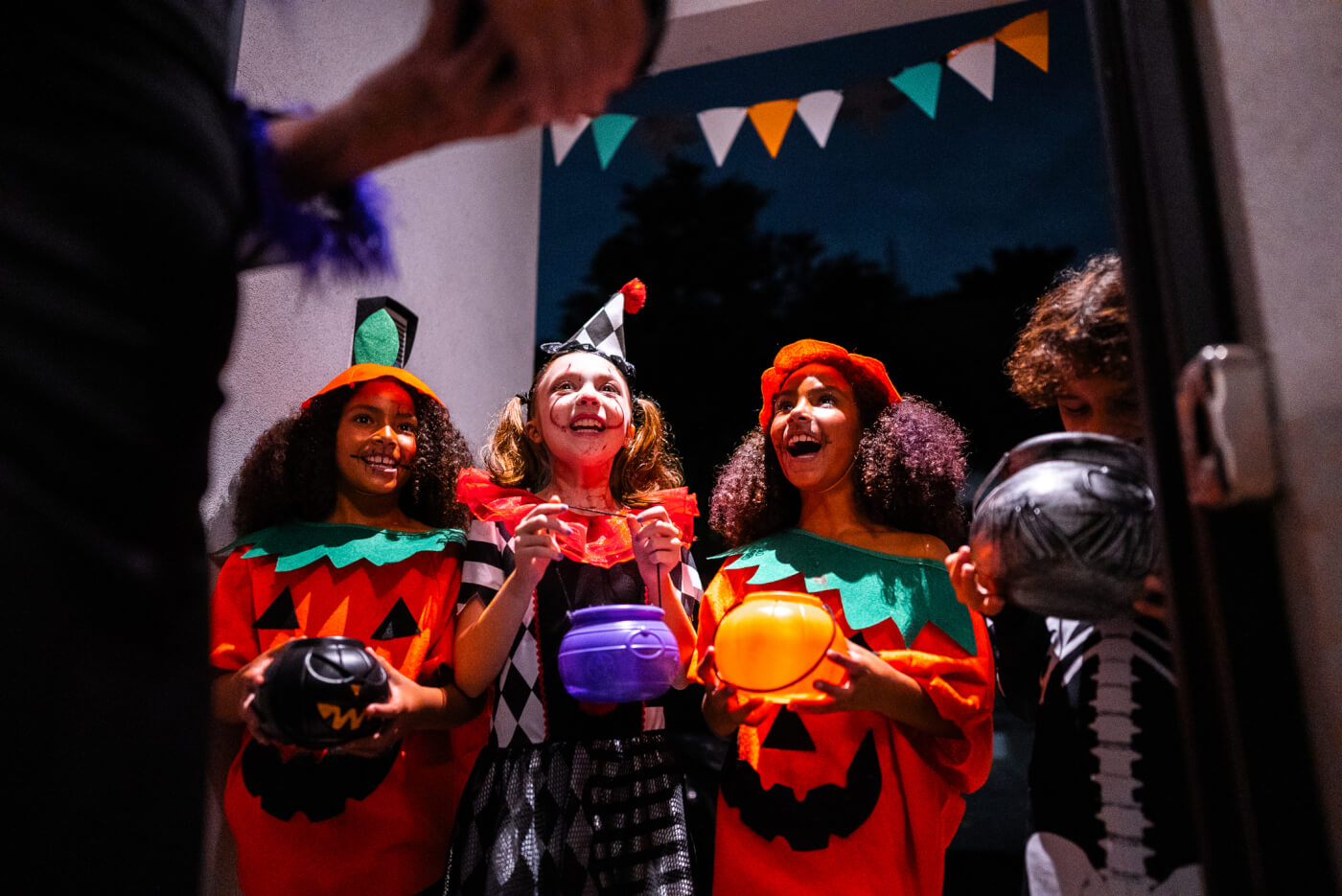 four kids dressed up for Halloween at a neighbor's door asking for candy. It is dark outside and two of the kids are dressed up as pumpkins, one kid is dressed up as a clown, and the other kid is dressed up as a skeleton. 
